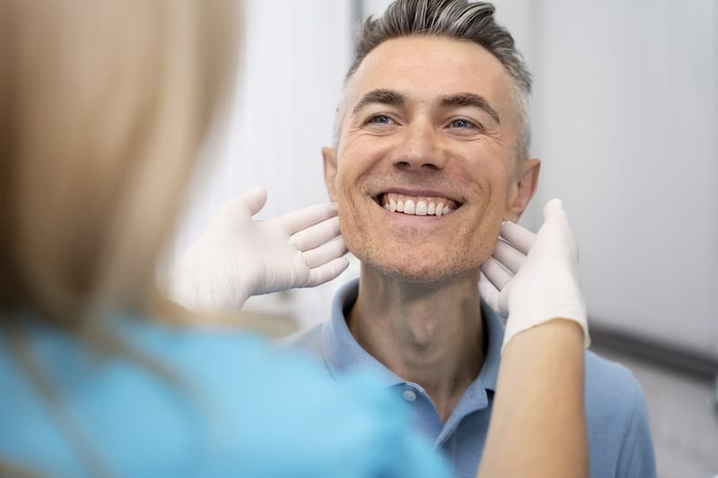 A smiling middle-aged man has his jaw examined by a dentist wearing gloves.