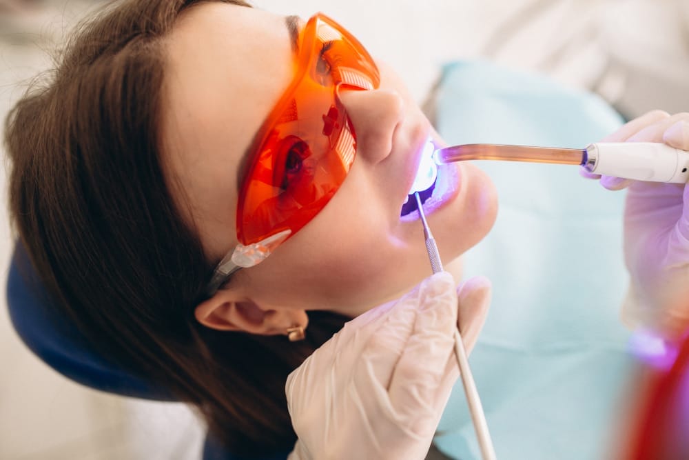 A woman receiving a dental procedure while wearing protective orange glasses. A dentist uses a curing light to harden a material inside her mouth.